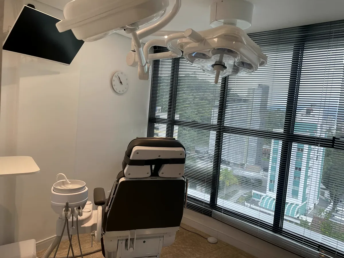 A room at the clinic in Blumenau: a dental chair under a two-headed surgical light, a screen on the wall, and a wide window with blinds looking out over the city