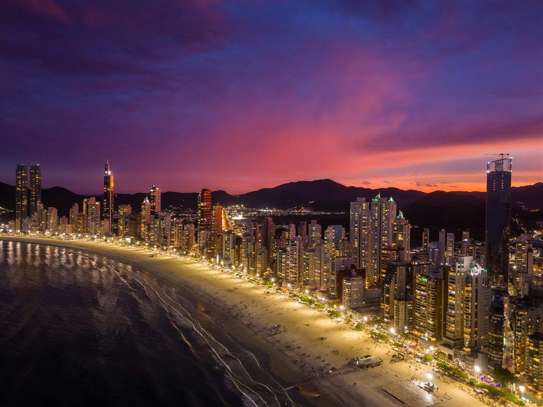 Balneário Camboriú at night from above: the curving beach, the lit beachfront and the high-rises under a red sky