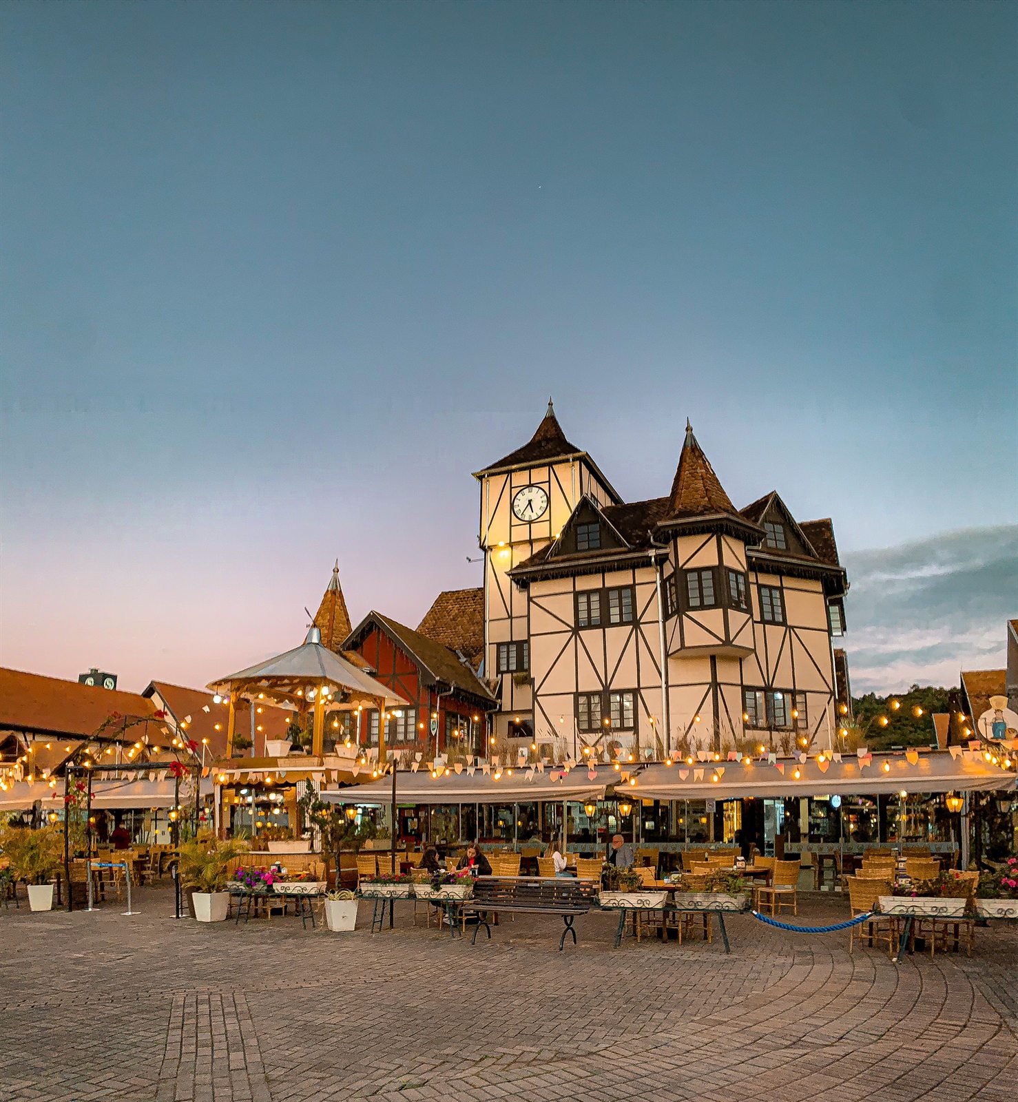 The Parque Vila Germânica at dusk: half-timbered pavilions with a clock tower, string lights and outdoor tables