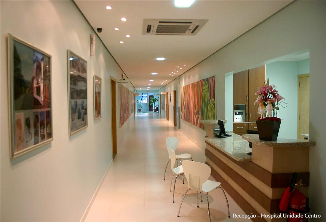 A reception desk with a vase of pink lilies at the start of a long, bright corridor hung with framed pictures, with white chairs beside the desk; in the bottom corner, the caption “Recepção – Hospital Unidade Centro”