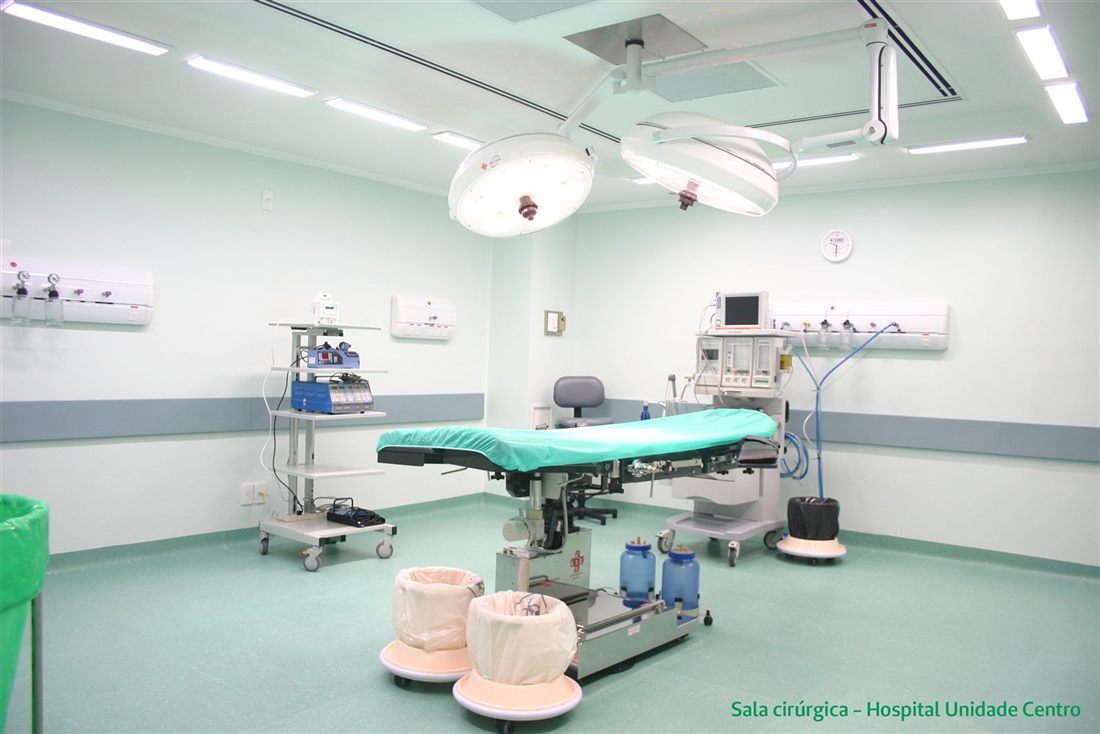 An empty operating room: two ceiling-mounted surgical lights above the operating table, an anesthesia machine with its monitor at the back and an equipment cart on the left; in the bottom corner, the caption “Sala cirúrgica – Hospital Unidade Centro”