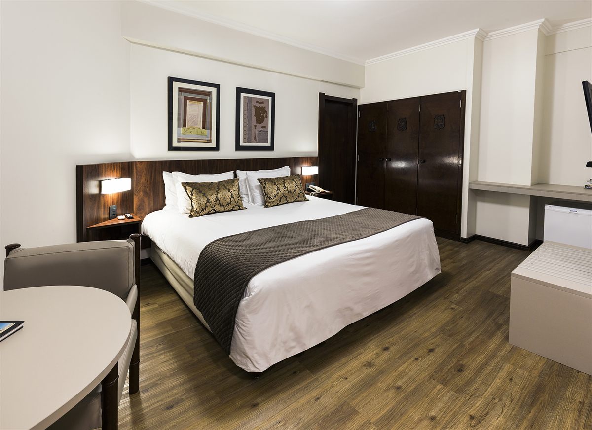 A suite at the Plaza Blumenau: a king bed with a wooden headboard and a round table by the window