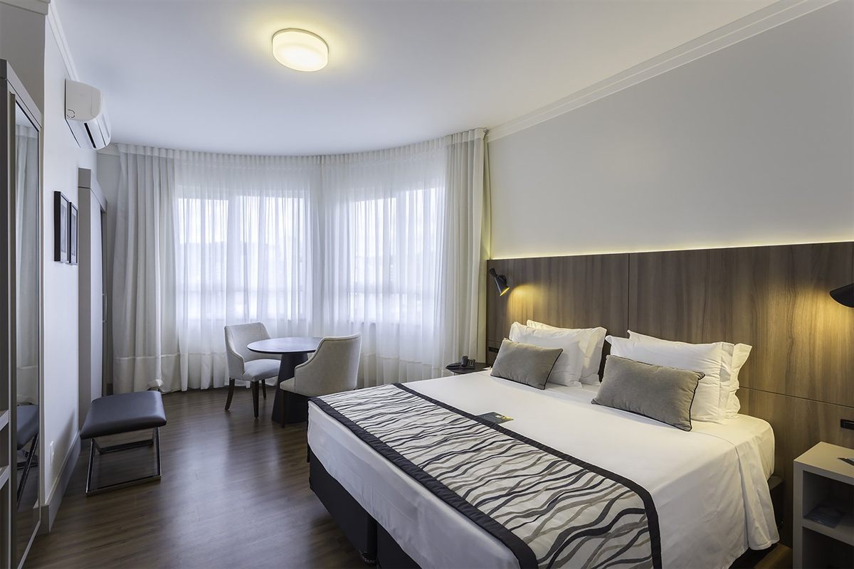 A room at the Slaviero in Blumenau: a king bed, a small round table and a curved window onto the city
